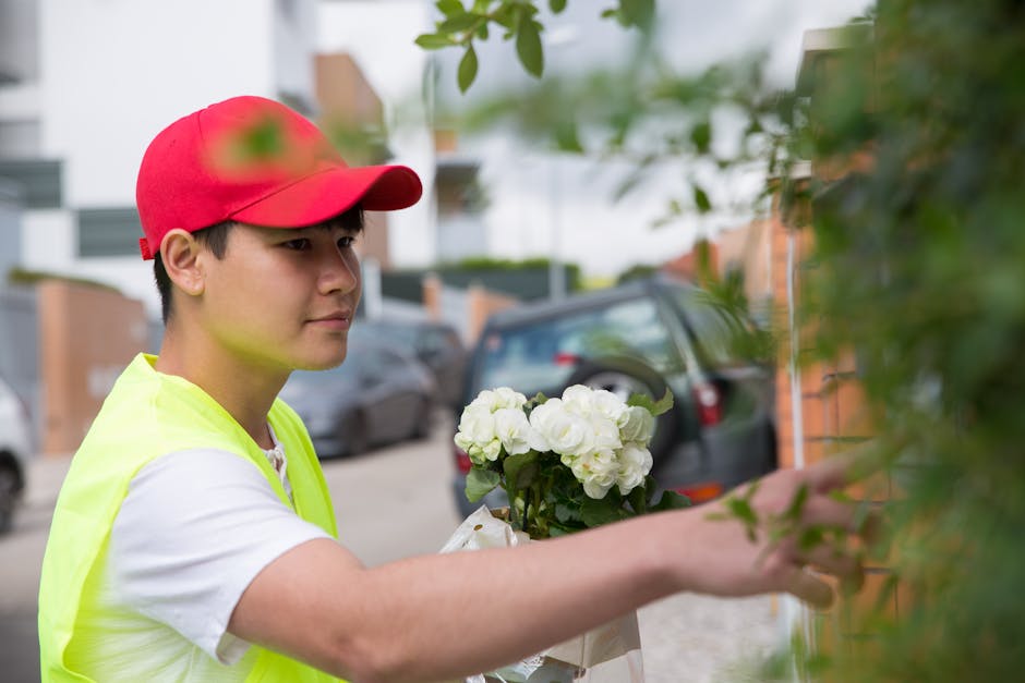 A young male florist delivering fresh white hydrangeas wrapped in white paper and tied with a ribbon. He is wearing a bright yellow high-visibility vest and a red cap, standing outdoors near a residen