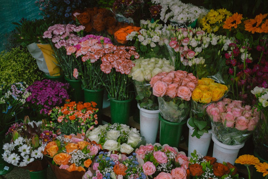 A vibrant display of fresh flowers arranged in various bouquets and individual stems, including pink roses, yellow roses, white roses, orange carnations, purple chrysanthemums, white daisies, and pink