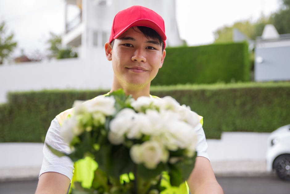 A smiling delivery person wearing a red cap and a high-visibility yellow vest holds a basket of fresh, white flowers with green foliage, ready for flower delivery outside a residential area. The bouqu