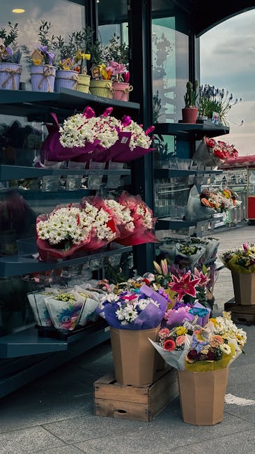 A man dressed in a red jacket and matching red cap is holding a floral gift wrapped in colorful, geometric-patterned paper featuring yellow, orange, blue, and pink triangles. The bouquet contains a va