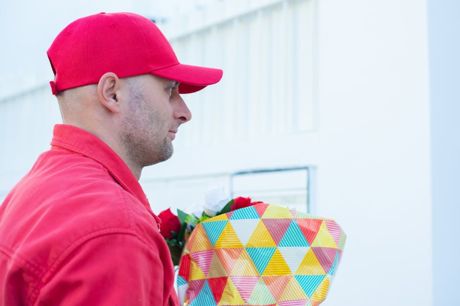 A man dressed in a red cap and red jacket holding a bouquet of fresh flowers wrapped in colorful geometric-patterned paper, with vibrant red, white, and green blooms visible. He is standing outdoors i